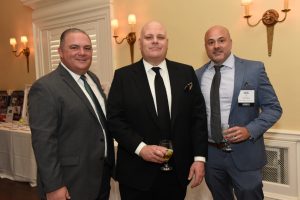 Three men in suits posing together at a formal event, each holding a glass of wine.