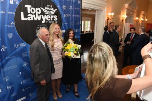 Three people pose for a photo at the 'Top Lawyers of Long Island' event, with a woman in beige dress holding a bouquet in front of a blue sponsor banner.