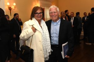 Smiling older couple posing for a photo at an indoor networking event; woman in a cream coat and scarf with a black handbag, man in a blue shirt and blazer.