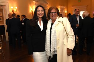 Two women pose for a photo at a formal reception, one in a black blazer and white dress, the other in a cream coat and white scarf, smiling in a warmly lit room.