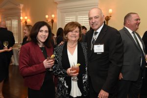 Three attendees at a reception pose for the camera, two women and a man in a suit, each holding a cocktail with lemon wedges.