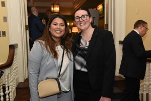 Two women in business attire smiling at the camera at a social event in a hallway with other guests in the background.
