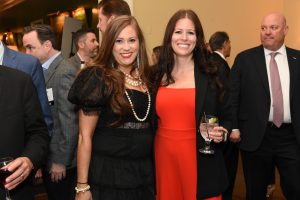 Two smiling women in cocktail attire pose for a photo at a social event; one wears a black lace dress with pearl necklace, the other a red dress with a black blazer holding a drink.
