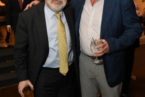 Two men in suits posing with drinks at a formal event, smiling for the camera.",