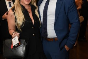 Two well-dressed attendees posing for a photo at a formal event: a blonde woman in a black outfit and a man in a blue suit.