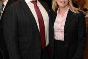 Two professionals pose for a photo at a formal event: a man in a pinstripe suit with a red tie and a white shirt, and a woman in a black blazer over a pink striped blouse.