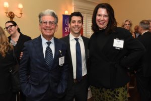 Three people in business attire pose for a photo at a formal event, smiling at the camera with name badges visible on their jackets on a warm-lit background.