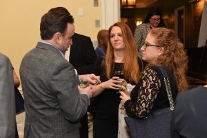 Three professionals converse at a cocktail reception, each holding a drink in a hallway setting.