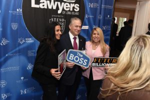 Three people pose at a lawyers event; the man in the center holds a 'The BOSS' sign, flanked by two women holding signs.