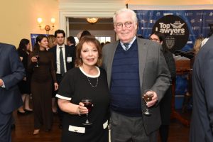 Smiling couple in formal attire posing for the camera at a reception, each holding a glass of wine, with guests mingling in the background and a 'Top Lawyers' banner visible.