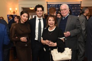 Four adults at a formal reception posing for a photo; woman in a brown dress, man in a dark suit, and an older couple with a beige handbag, all holding wine glasses.