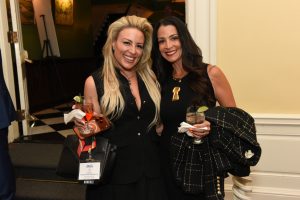Two women in black outfits pose together at a formal event, each holding a cocktail with a lime wedge nearby.