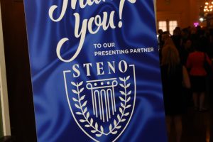 Blue event sign with white 'Thank you!' text and crest, thanking presenting partner STENO, placed on a stand near a doorway in a crowded lobby.