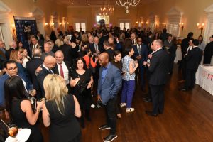 Crowded formal networking event in a chandeliers-lit ballroom with people mingling and holding drinks in business attire.