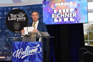 Man in a blue suit speaks at a podium during a legal event, with a 'Welcome' banner and 'Top Lawyers of Long Island' backdrop behind him.