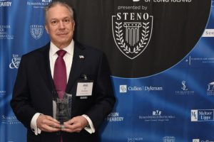Man in a dark suit and burgundy tie holding a glass award at the 2026 Top Lawyers of Long Island event, in front of a blue sponsor backdrop.