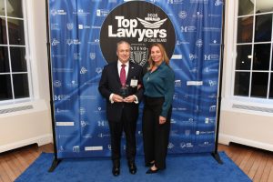 Man in a dark suit and red tie holds a glass award beside a woman in a teal blouse at a 'Top Lawyers of Long Island' backdrop.