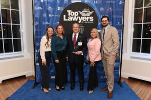 Group of five adults posing on a blue carpet at a lawyers awards event; one man center holds a trophy, backdrop reads 'Top Lawyers of Long Island' with logos on blue background