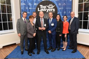 Eight professionals in formal attire pose on a blue carpet in front of a 'Top Lawyers of Long Island 2026' backdrop; the man in the center holds a glass award.
