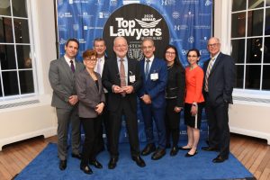 Eight professionals in business attire pose on a blue carpet in front of a 'Top Lawyers' backdrop, holding a glass award at an awards ceremony.