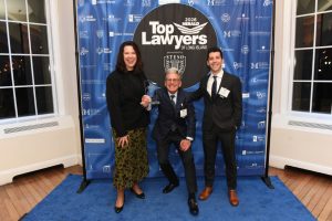 Three well-dressed individuals pose on a blue carpet at a Top Lawyers of Long Island event, with a trophy in the center.