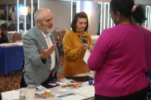 Man in a light-blue blazer talks to a woman in a mustard sweater at a booth; papers and candy are spread across the table, with a person in pink in the foreground.