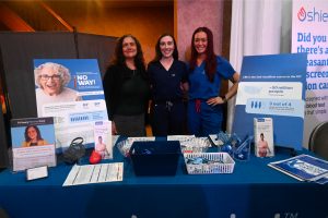 Three women in scrubs stand behind a blue information booth at a health fair, smiling with large posters about cancer screening nearby.