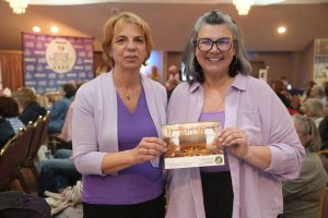 Two women stand side by side at an indoor expo, smiling, wearing lavender outfits and holding a brochure.