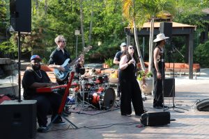 Outdoor band performance featuring keyboardist, bassist, drummer, and two vocalists in a sunny garden setting.