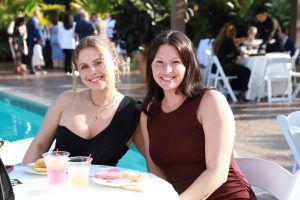 Two women smile at the camera while seated at a poolside table during an outdoor party, with drinks and fruit in front of them.