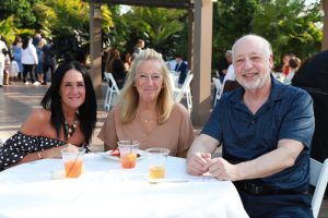 Three smiling adults seated at a white-clothed outdoor event table with colorful drinks in clear cups, posing for the photo with greenery and guests in the background.