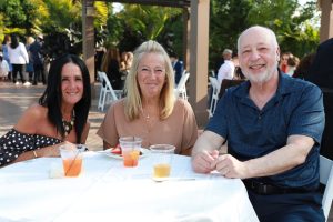 Three adults sit at a round table outdoors at a social event, smiling at the camera with drinks in plastic cups nearby.