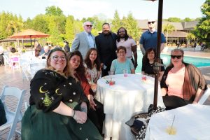 Group of nine people posing around a round poolside table at a sunny outdoor party.