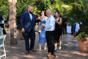 Group of adults in semi-formal attire chatting and toasting with drinks at an outdoor garden party with palm plants nearby.