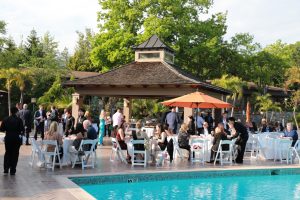 Outdoor poolside reception under a wooden pavilion; guests dine at round tables with white chairs and an orange umbrella by a sunny garden venue.