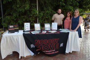 Three adults stand behind an information table outdoors, with a black 'Nobody Move' banner and flyers on display.