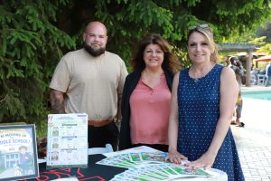 Three adults stand behind a table with school program brochures at an outdoor event near a pool and green trees.