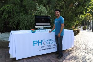 Man standing at a branded table with a desktop computer, outdoor garden backdrop, wearing a blue PH Consulting & Media shirt; table covered with a white cloth displaying the company logo.