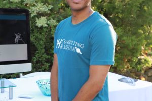 Smiling man in teal T-shirt standing outdoors beside a table with a computer, promoting consulting and media services.