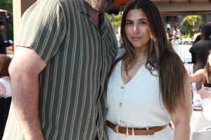 Couple posing together at an outdoor event, man in striped shirt and beard, woman in white belted outfit, smiling at camera.