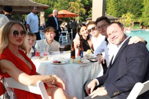 Group of seven adults around a round table by a pool at an outdoor party, wearing semi-formal attire and enjoying drinks and snacks.