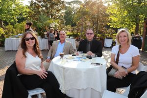 Four adults sit at a round table outdoors during a social event, smiling at the camera with drinks and plates in front of them.