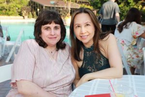 Two women smile at a poolside outdoor event, seated at a table with drinks in a sunny setting.