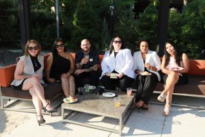 Six adults sitting on an outdoor sofa with plates of food and drinks on a table in front of them.