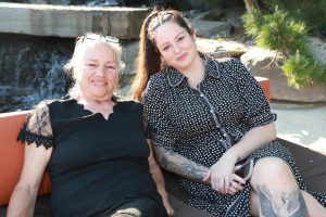 Two smiling women sit on an outdoor couch by a water feature, one in a black lace-edged dress and the other in a black-and-white polka-dot dress.