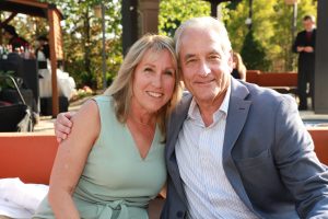 Smiling older couple posing together outdoors at a sunny event, arm around each other on a bench.
