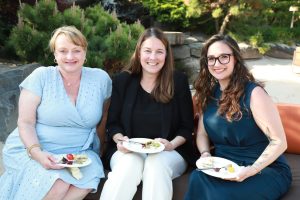 Three women sit on a bench outdoors, smiling, each with a small plate of appetizers in a sunny garden setting.