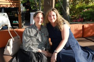 Two women sit closely on an orange outdoor sofa, smiling at the camera at a garden party; one wears a black-and-white houndstooth blouse and dark skirt, the other a navy blue sparkly dress with blond wavy hair next to her. A beige handbag rests beside them on the sofa.