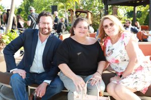 Three adults sitting on a park-side bench at an outdoor event, smiling at the camera.