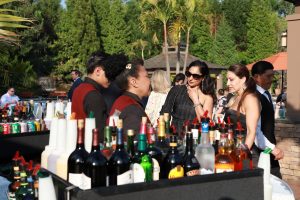 Group of well-dressed guests chat with bartenders at an outdoor cocktail bar, bottles and mixers lined up on the counter.